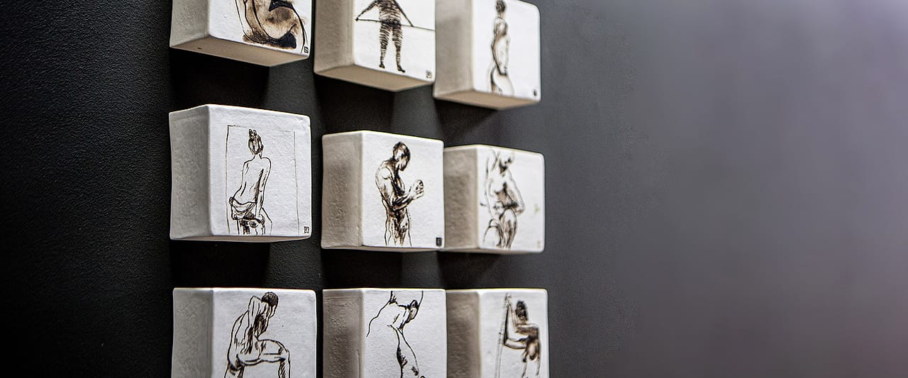 Figure studies drawn on a grid of white ceramic blocks mounted on a dark wall in the practice.
