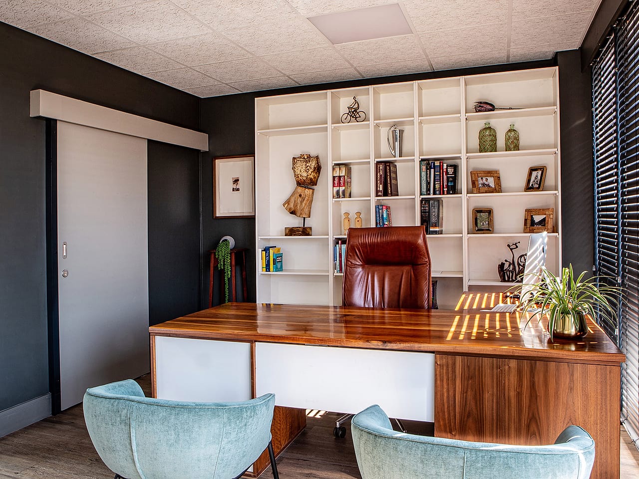 A consulting room with a walnut desk, two soft green armchairs and a wall of books.