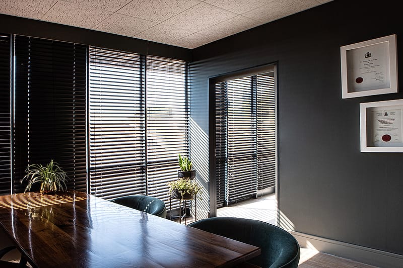 A consulting room table beside a window with timber blinds and a potted plant.