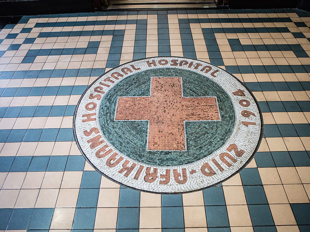 The original 1904 mosaic floor at Zuid-Afrikaans Hospital, a red cross set into tiled lettering reading “Zuid-Afrikaans Hospitaal”.