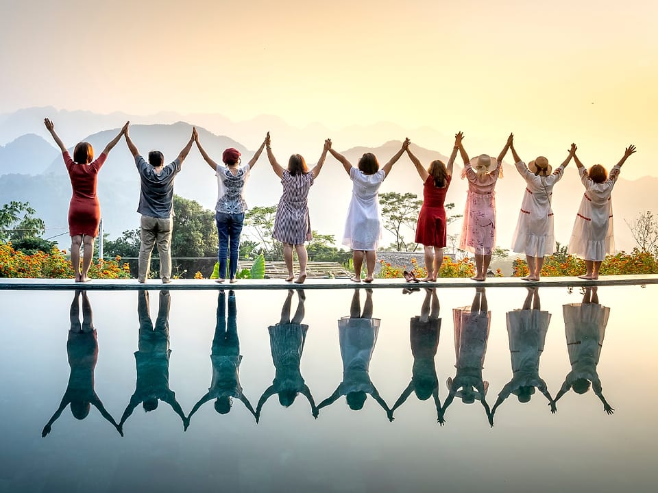 A line of people standing with arms raised at sunset, reflected in still water.